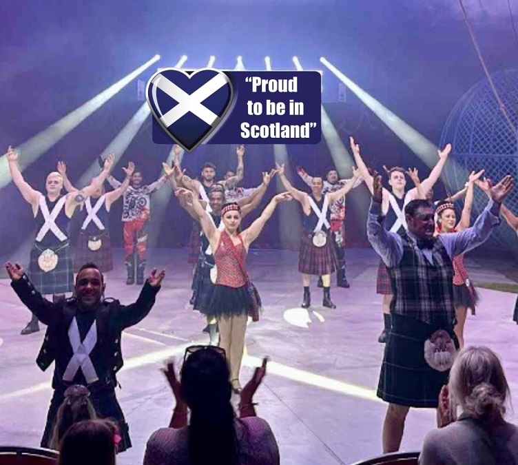 Stage performance with kilted dancers raising their arms in unison, a Scottish flag emblem and the text “Proud to be in Scotland” displayed above, while audience members watch and applaud in the foreground.