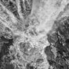 Black-and-white aerial view of rushing water cascading over dark rocky cliffs, creating swirling foam and dramatic textures.
