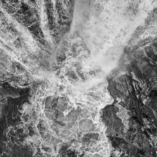 Black-and-white aerial view of rushing water cascading over dark rocky cliffs, creating swirling foam and dramatic textures.
