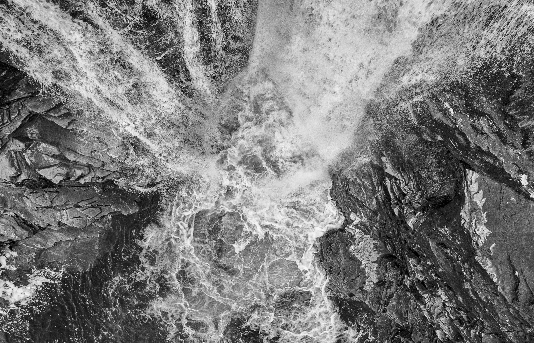 Black-and-white aerial view of rushing water cascading over dark rocky cliffs, creating swirling foam and dramatic textures.