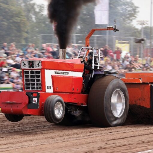 Red tractor pulling a weighted sled during a tractor-pulling competition on a dirt track, with black smoke billowing from the exhaust and spectators watching in the background.