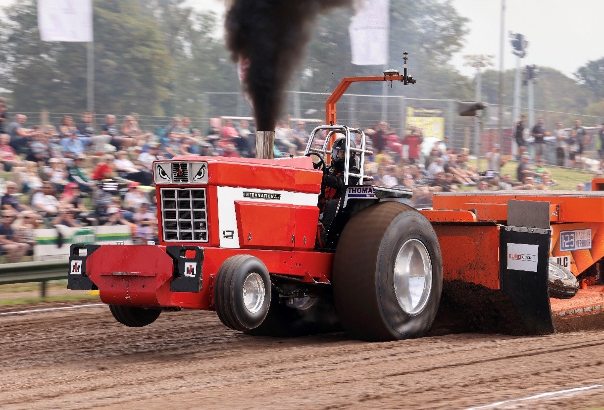 Red tractor pulling a weighted sled during a tractor-pulling competition on a dirt track, with black smoke billowing from the exhaust and spectators watching in the background.