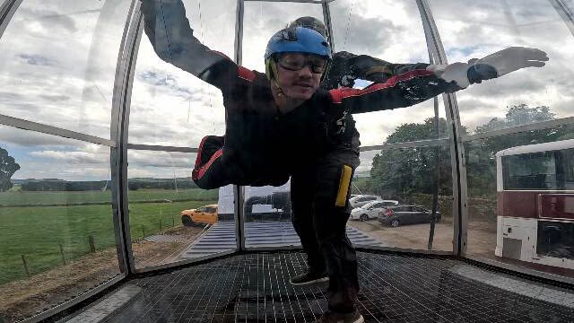 Person wearing a helmet and flight suit experiencing an indoor skydiving session in a vertical wind tunnel, hovering above a grated floor with countryside visible through the transparent walls.