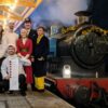Group of people in festive costumes standing on a decorated railway platform beside a steam train at night, holding menu boards under illuminated station lights.