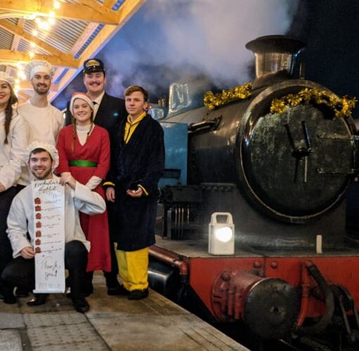 Group of people in festive costumes standing on a decorated railway platform beside a steam train at night, holding menu boards under illuminated station lights.