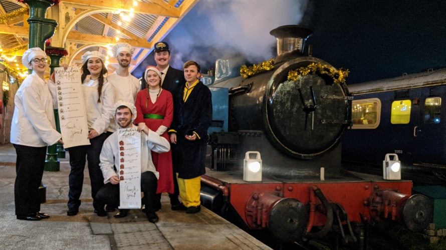Group of people in festive costumes standing on a decorated railway platform beside a steam train at night, holding menu boards under illuminated station lights.