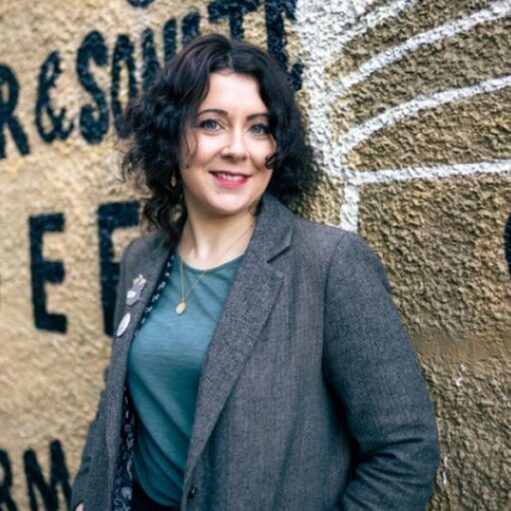 Portrait of a person wearing a grey blazer and teal top, standing in front of a textured wall with large painted lettering and mural artwork in the background.