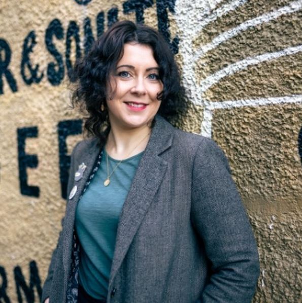 Portrait of a person wearing a grey blazer and teal top, standing in front of a textured wall with large painted lettering and mural artwork in the background.