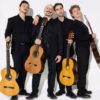 Four musicians dressed in black standing with classical guitars against a plain light background, posing together for a promotional group portrait.