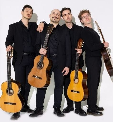 Four musicians dressed in black standing with classical guitars against a plain light background, posing together for a promotional group portrait.