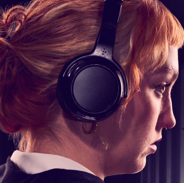 Close-up side-profile portrait of a person with red hair wearing large black over-ear headphones, against a dark background with soft lighting highlighting the face and headphones.