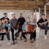 Seven-piece folk band seated and standing in a spacious industrial-style room, holding a variety of instruments including accordion, fiddle, acoustic guitar, bass guitar and traditional stringed instruments, with exposed brick walls and large windows in the background.