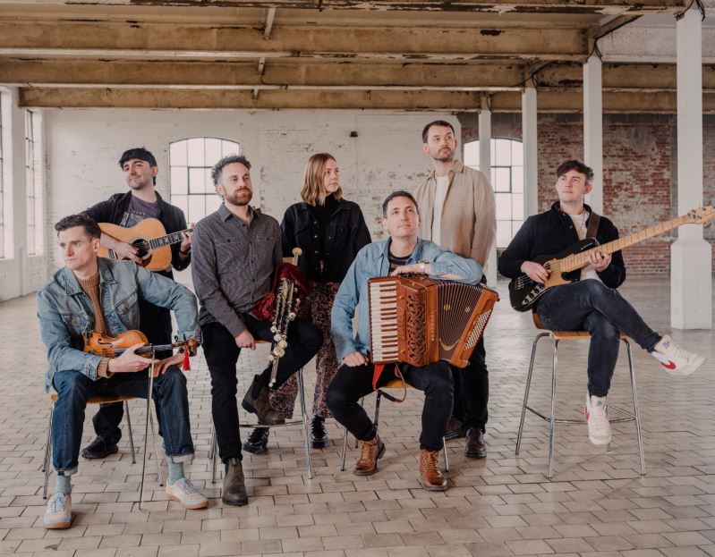 Seven-piece folk band seated and standing in a spacious industrial-style room, holding a variety of instruments including accordion, fiddle, acoustic guitar, bass guitar and traditional stringed instruments, with exposed brick walls and large windows in the background.
