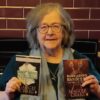 Person with grey hair and glasses holding two books by Maggie Craig in front of a tiled wall, including Damn’ Rebel Bitches: The Women of the ’45 and Bare-Arsed Banditti: The Men of the ’45.