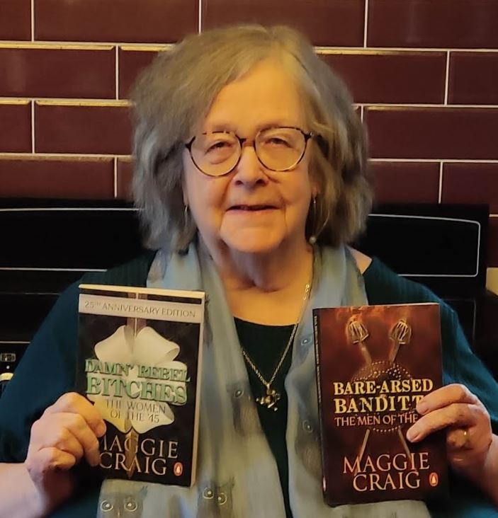 Person with grey hair and glasses holding two books by Maggie Craig in front of a tiled wall, including Damn’ Rebel Bitches: The Women of the ’45 and Bare-Arsed Banditti: The Men of the ’45.