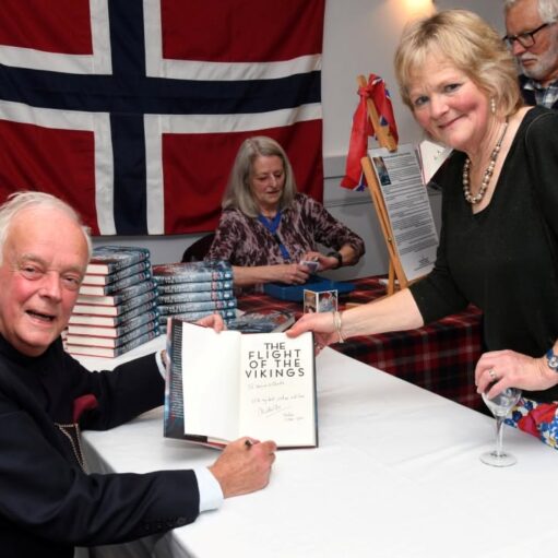 Author signing a copy of The Flight of the Vikings at a book event, seated at a table with a guest holding the open book, while stacks of books and a large Norwegian flag are visible in the background.