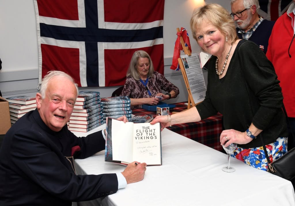 Author signing a copy of The Flight of the Vikings at a book event, seated at a table with a guest holding the open book, while stacks of books and a large Norwegian flag are visible in the background.