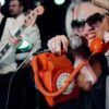 Performer wearing large sunglasses holds a bright orange rotary telephone and sings into a microphone on stage, while musicians with guitars play in the background under colourful concert lighting.