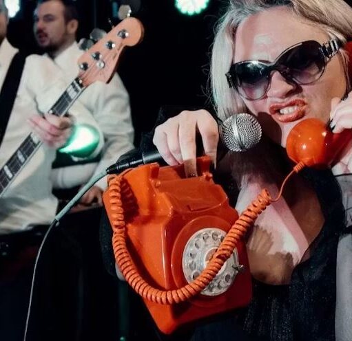 Performer wearing large sunglasses holds a bright orange rotary telephone and sings into a microphone on stage, while musicians with guitars play in the background under colourful concert lighting.