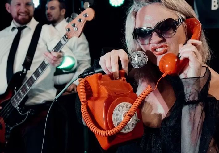 Performer wearing large sunglasses holds a bright orange rotary telephone and sings into a microphone on stage, while musicians with guitars play in the background under colourful concert lighting.
