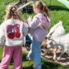 Two children standing beside a wire fence at a petting farm, watching several goats in a grassy enclosure, including a white horned goat in the foreground, with play equipment visible in the background.
