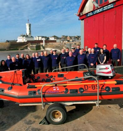 Group photograph of RNLI volunteers standing outside a lifeboat station beside an orange inflatable rescue boat, with a harbour, beach and white lighthouse visible in the background.