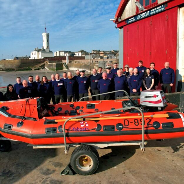 Group photograph of RNLI volunteers standing outside a lifeboat station beside an orange inflatable rescue boat, with a harbour, beach and white lighthouse visible in the background.