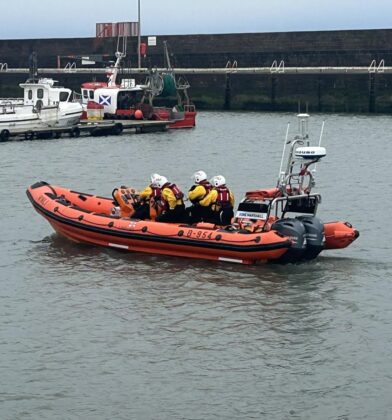 Group photograph of RNLI volunteers standing outside a lifeboat station beside an orange inflatable rescue boat, with a harbour, beach and white lighthouse visible in the background.