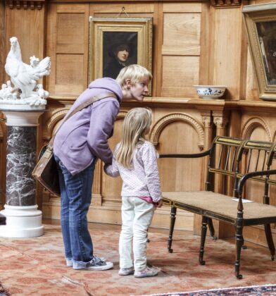 Visitor and child examining paintings, sculptures and historic furniture in an ornate wood-panelled room within a heritage house or museum.