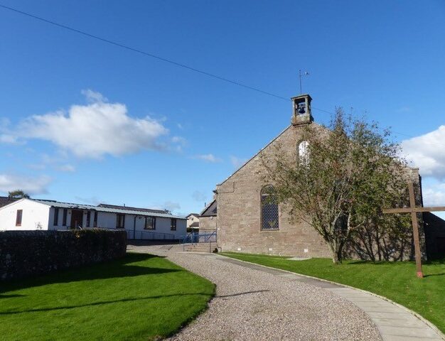 Stone church building in Letham with a bellcote and arched window, set beside a gravel pathway, grassy grounds and a wooden cross, under a bright blue sky with scattered clouds.