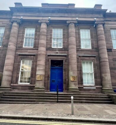 Historic sandstone civic building with tall classical columns, a central blue doorway and broad stone steps leading from the street, with a traditional red telephone box positioned nearby.