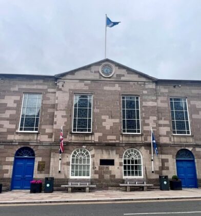 Symmetrical stone town hall building featuring a central clock, blue entrance doors, arched ground-floor windows and flags displayed on the front façade facing the main street.