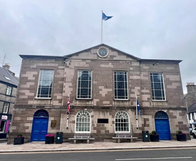 Symmetrical stone town hall building featuring a central clock, blue entrance doors, arched ground-floor windows and flags displayed on the front façade facing the main street.