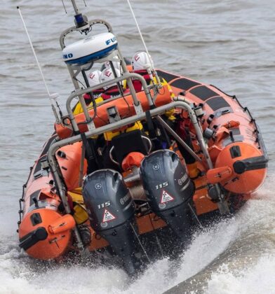 RNLI inshore lifeboat powering through rough water, with crew members in protective gear aboard and sea spray rising behind the twin outboard engines.