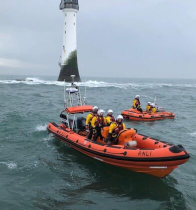 Two RNLI lifeboats carrying volunteer crew members on choppy seas near a tall white lighthouse, with waves breaking around the rocky shoreline in the background.