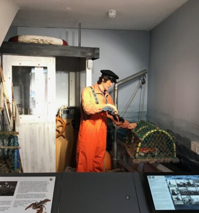 Museum display recreating a traditional fishing scene, featuring a life-sized fisherman mannequin in orange waterproof clothing beside lobster pots, fishing equipment and a small boat cabin, with interpretive panels in the foreground.