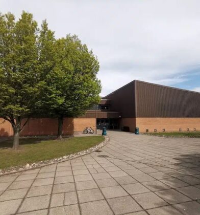 Modern sports centre building with brick and dark-panelled exterior, set around a paved forecourt with mature trees and landscaped grounds.
