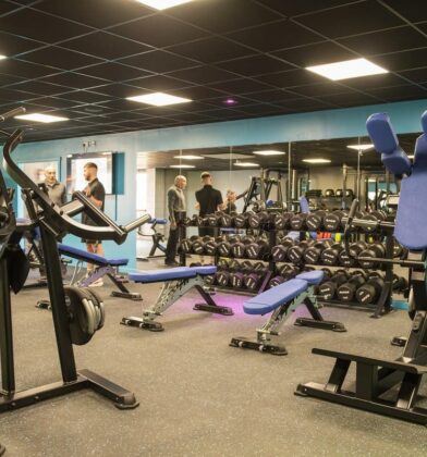 Well-equipped fitness gym featuring resistance machines, free weights, benches and mirrored walls, with people using the exercise facilities.