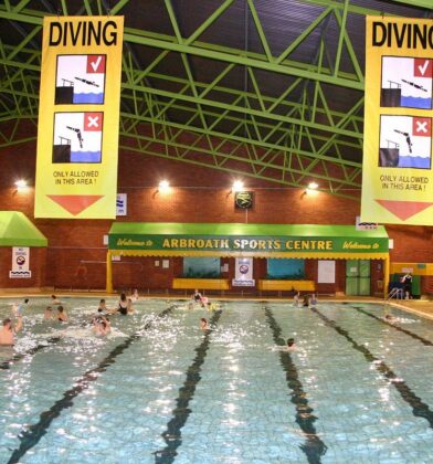 Indoor swimming pool at Arbroath Sports Centre, with people swimming and playing in marked lanes beneath large diving safety signs suspended from a high green steel-framed ceiling.