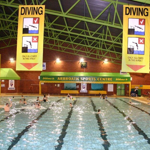 Indoor swimming pool at Arbroath Sports Centre, with people swimming and playing in marked lanes beneath large diving safety signs suspended from a high green steel-framed ceiling.