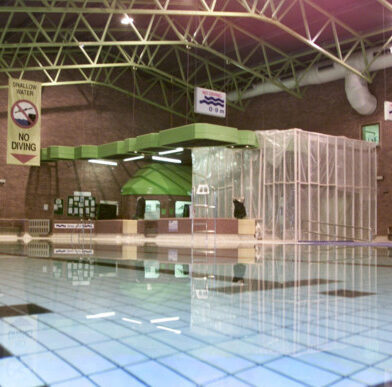 Wide view of the Arbroath Sports Centre swimming pool, showing calm water, poolside seating and diving safety signage within a spacious brick-lined leisure facility.