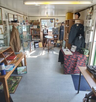 Museum exhibition room featuring display cases, historical artefacts, military uniforms, books, photographs and interpretive information panels arranged throughout a compact gallery space.