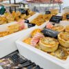 Market stall displaying a variety of freshly baked savoury pies and pastries in trays, with handwritten flavour signs and striped awnings visible in the background.