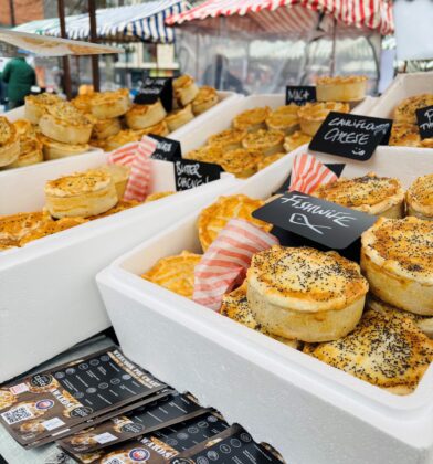 Market stall displaying a variety of freshly baked savoury pies and pastries in trays, with handwritten flavour signs and striped awnings visible in the background.