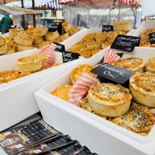 Market stall displaying a variety of freshly baked savoury pies and pastries in trays, with handwritten flavour signs and striped awnings visible in the background.