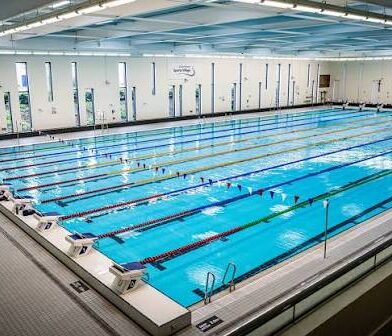 Large indoor swimming pool with multiple marked lanes, viewed from the spectator seating area, within a spacious modern leisure facility.