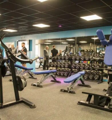Modern fitness gym featuring resistance machines, free weights, weight benches and mirrored walls, with people using exercise equipment throughout the spacious workout area.