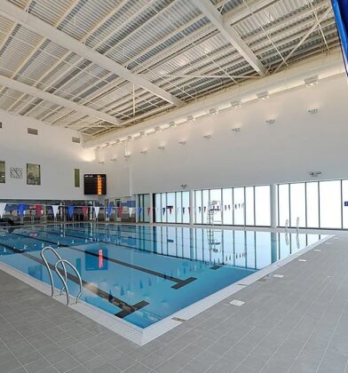 Bright indoor swimming pool with marked lanes, poolside seating and large frosted windows, housed within a modern leisure facility with high ceilings.