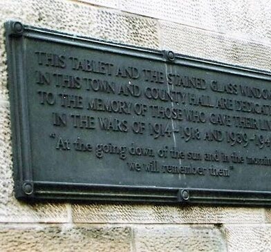 Bronze memorial plaque mounted on a stone wall, commemorating those who gave their lives in the First and Second World Wars, with an inscription ending, “At the going down of the sun and in the morning we will remember them.”