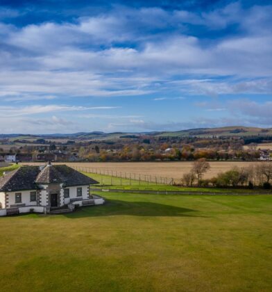 Small historic camera obscura building standing on a large open lawn, with panoramic views across surrounding farmland, rolling hills and woodland beneath a dramatic blue sky filled with scattered clouds.